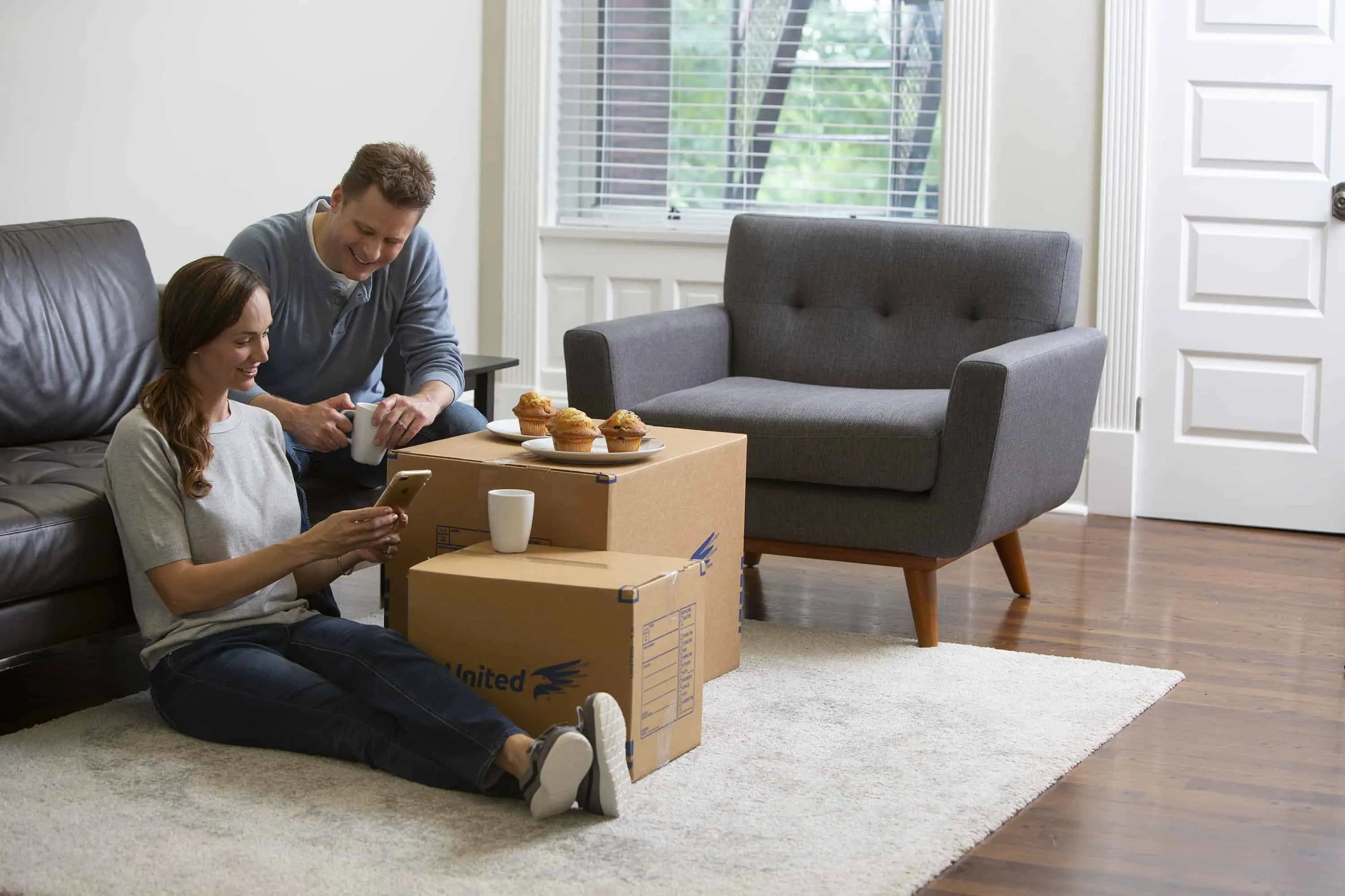 How to Pack the Living Room for Moving - young couple taking a break from packing their living room, using a box as a table for their breakfast muffins - United Van Lines